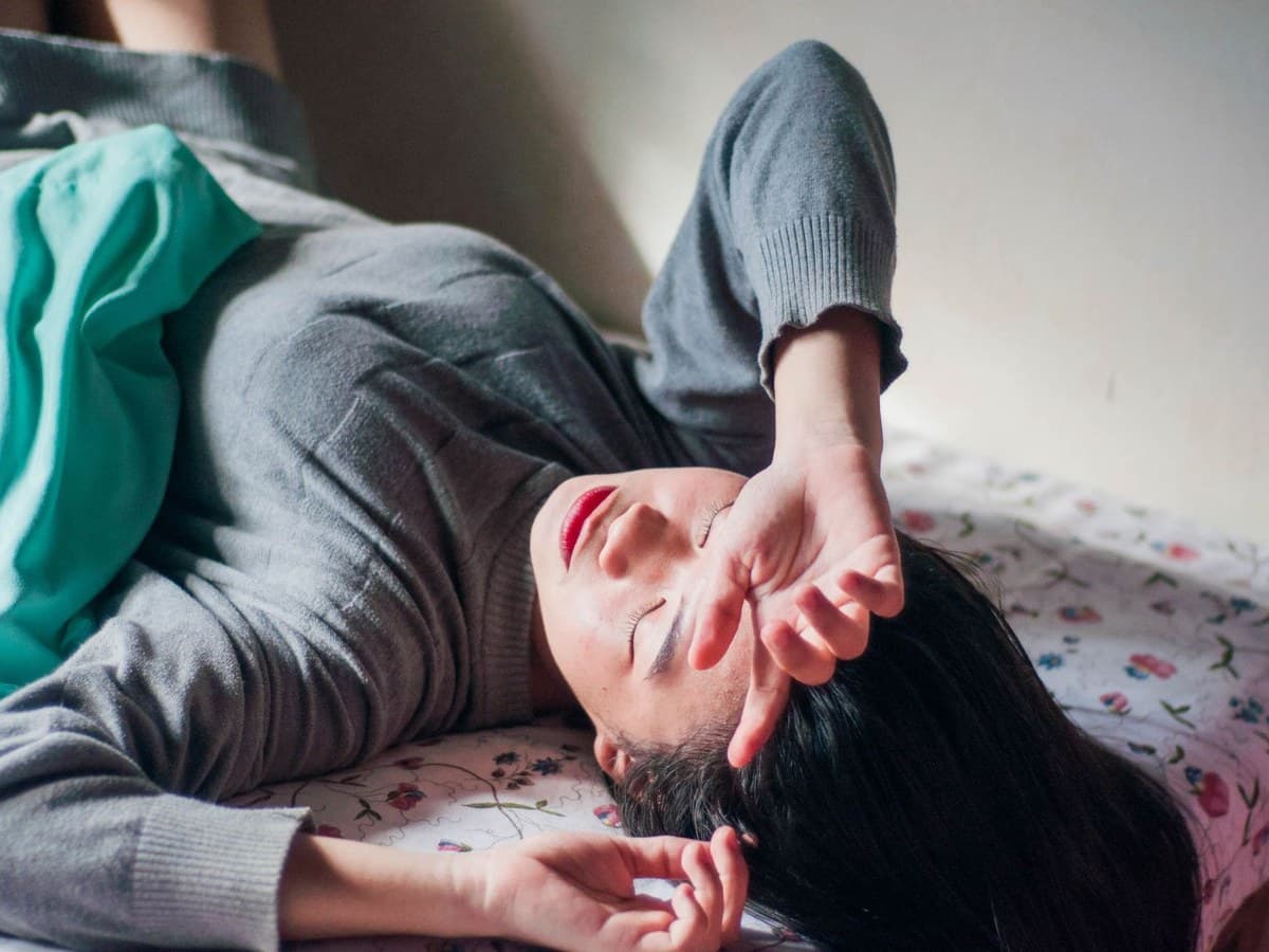 A woman lying down on a bed with an arm across her forehead in soft daylight