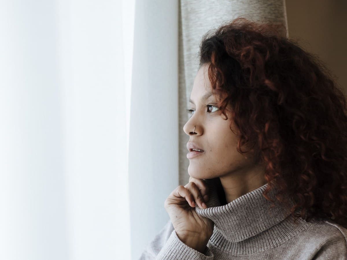A woman looking out of a window in soft daylight, hand resting near her chin